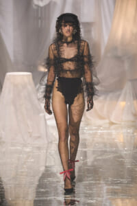 Model on a runway wearing a sheer black lace bodysuit with veil, gloves, and black heels with red bow ankle ties.