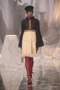 Model walking runway in sheer black top with gold embroidered buttons and cream skirt with fur trim, red lace tights, black hat with veil, and red bow-heeled shoes.