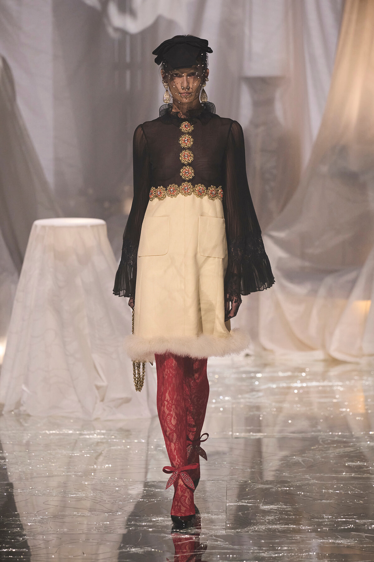 Model walking runway in sheer black top with gold embroidered buttons and cream skirt with fur trim, red lace tights, black hat with veil, and red bow-heeled shoes.