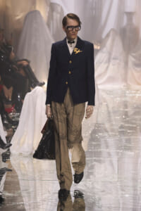 Male model walking the runway in a navy blazer with gold buttons, beige pleated pants, lace gloves, oversized glasses, and a large black handbag.
