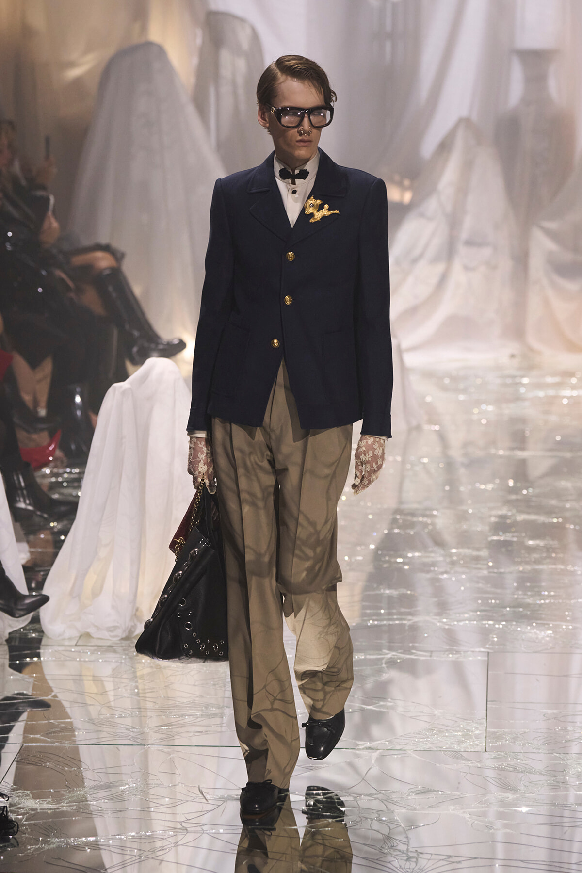 Male model walking the runway in a navy blazer with gold buttons, beige pleated pants, lace gloves, oversized glasses, and a large black handbag.