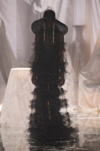 Model in a black, beaded, tiered gown with an oversized wide-brim hat and chandelier earrings, posing against draped white fabric.