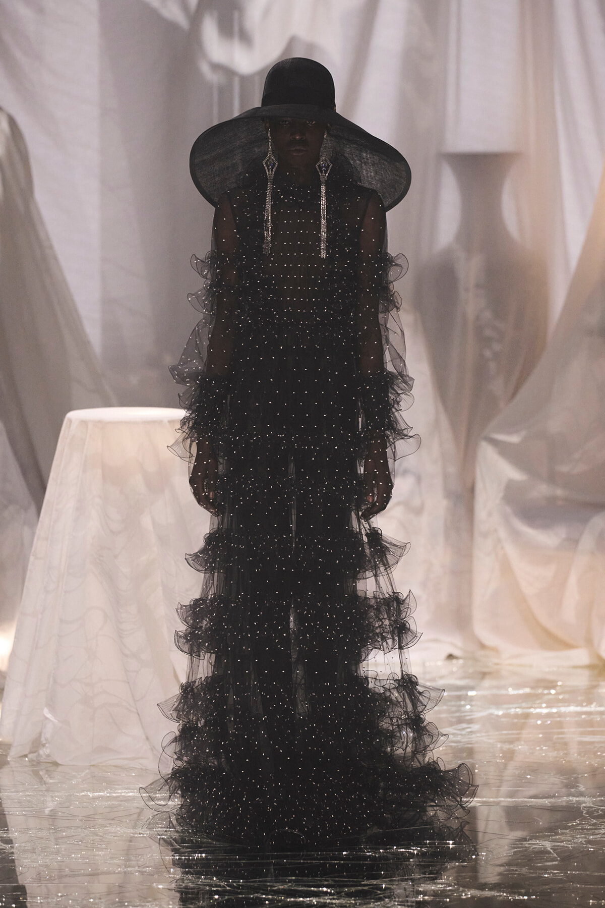 Model in a black, beaded, tiered gown with an oversized wide-brim hat and chandelier earrings, posing against draped white fabric.