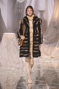 Model on a runway wearing a black-and-beige striped fur coat over a cream lace dress, carrying a tan shoulder bag.