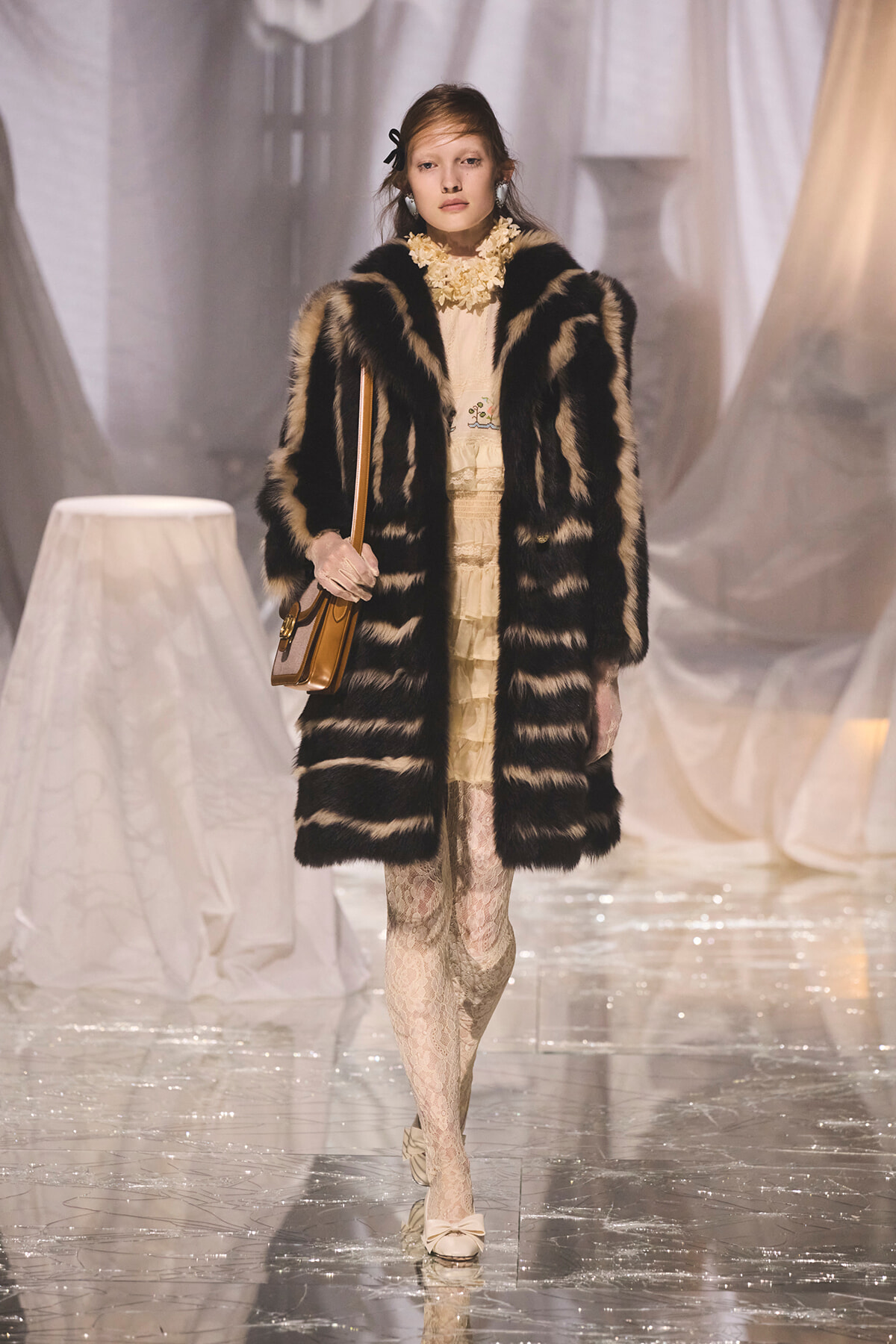 Model on a runway wearing a black-and-beige striped fur coat over a cream lace dress, carrying a tan shoulder bag.