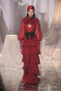 Model walking a red, layered ruffle gown with a black bow belt on a glossy runway.