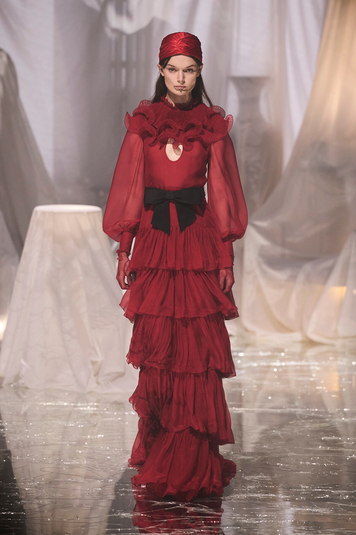 Model walking a red, layered ruffle gown with a black bow belt on a glossy runway.