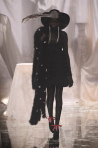 Model in a black lace dress with a long fur stole and a wide-brimmed feathered hat on a glossy runway, bold and dramatic.