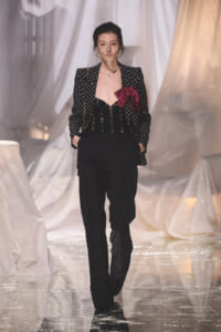 Model walking a fashion show wearing a black polka-dot blazer over a laced bodice with a large red bow, and black wide-leg pants.