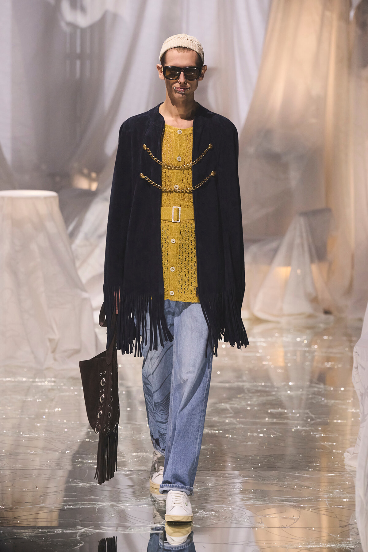 Model on runway wearing a mustard knit top under a fringed navy coat with gold chain details, light blue jeans, white sneakers, and a beige knit cap, carrying a brown fringe bag.