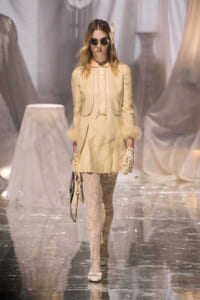 Runway model in a pale cream vintage-inspired coat-dress with lace tights, polka-dot gloves, and round sunglasses.