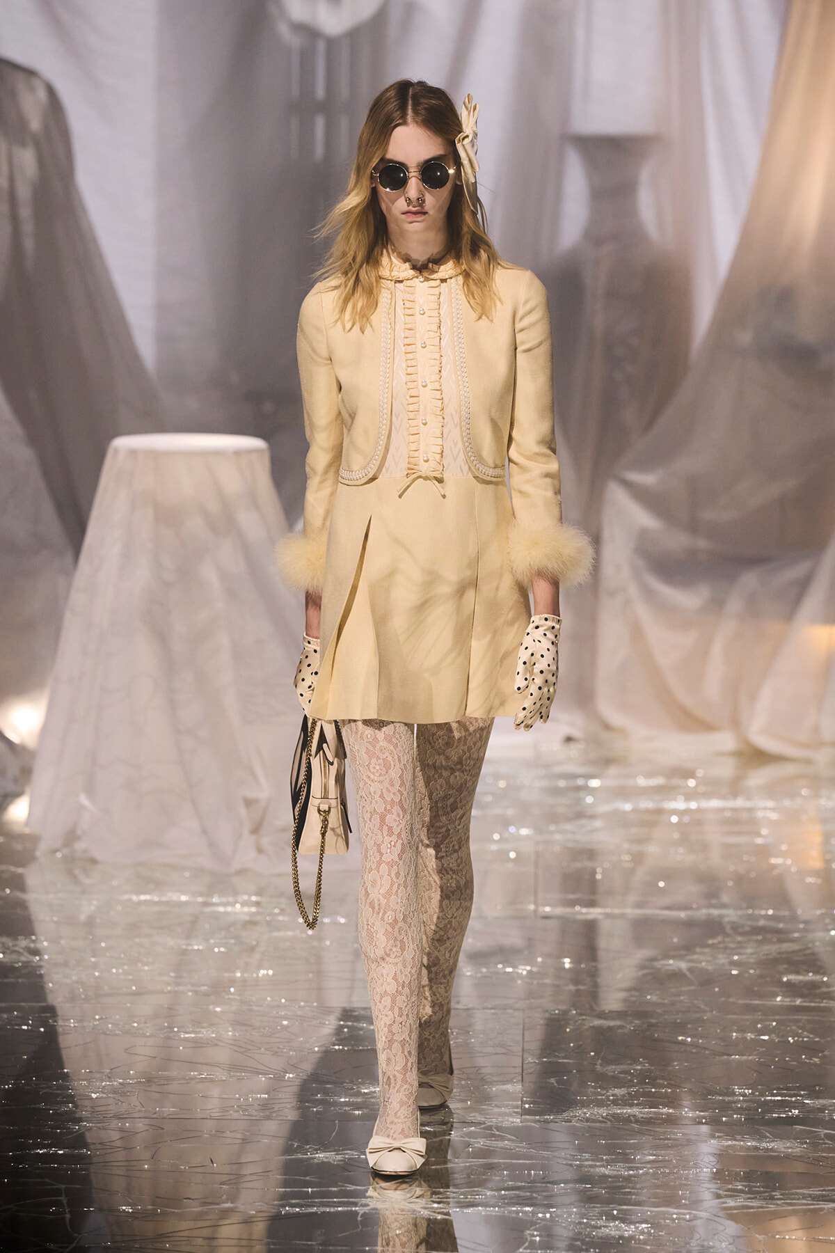 Runway model in a pale cream vintage-inspired coat-dress with lace tights, polka-dot gloves, and round sunglasses.