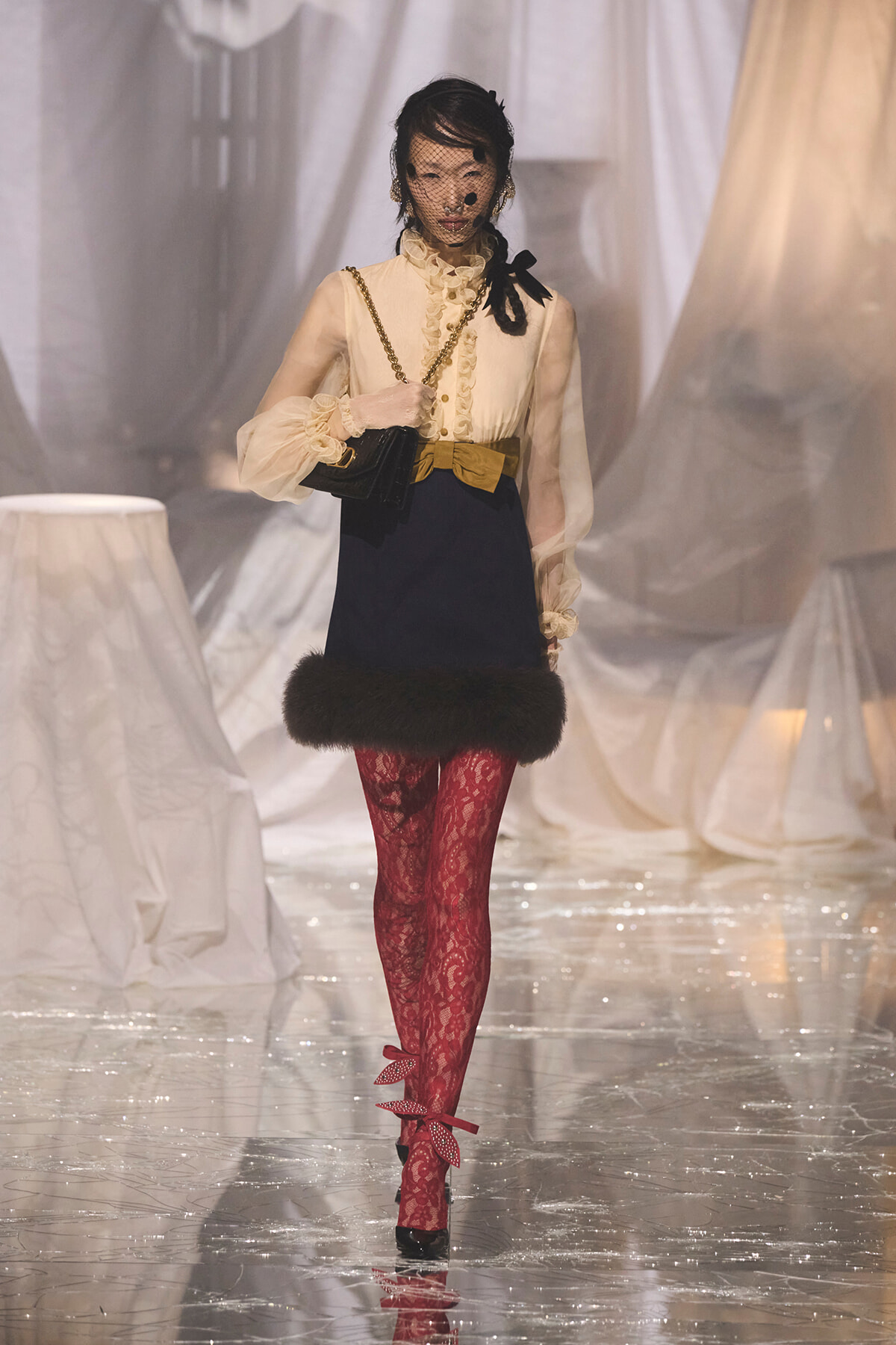 Model walking the runway in a vintage-inspired cream blouse with ruffles and sheer sleeves, navy skirt with a fur hem, a mustard bow belt, red lace tights, black heels, and a black chain-strap handbag (with netted veil over face).