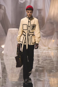 Model on a glossy runway wearing a cream belted jacket with black piping and patches, red knit cap, black trousers, and a patterned handbag.