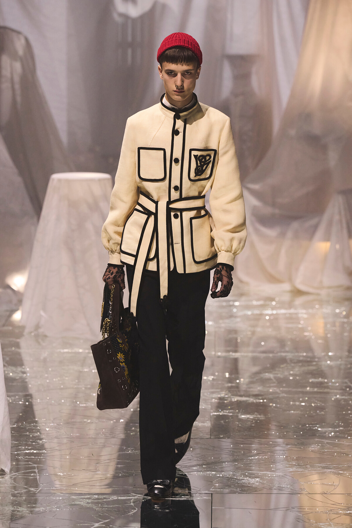 Model on a glossy runway wearing a cream belted jacket with black piping and patches, red knit cap, black trousers, and a patterned handbag.