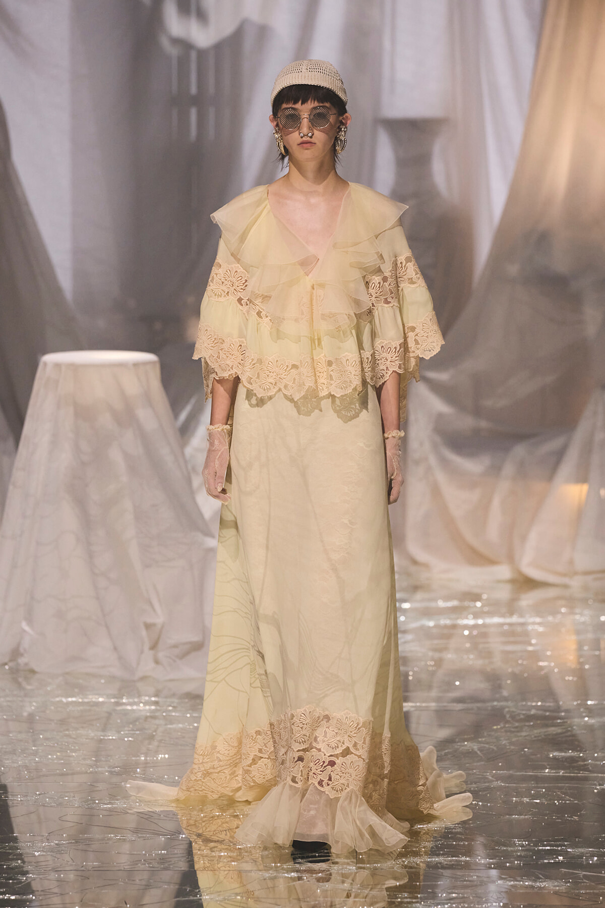 Model walks the runway in a pale cream gown with layered ruffles and lace trim, plus lace gloves and oversized round sunglasses.