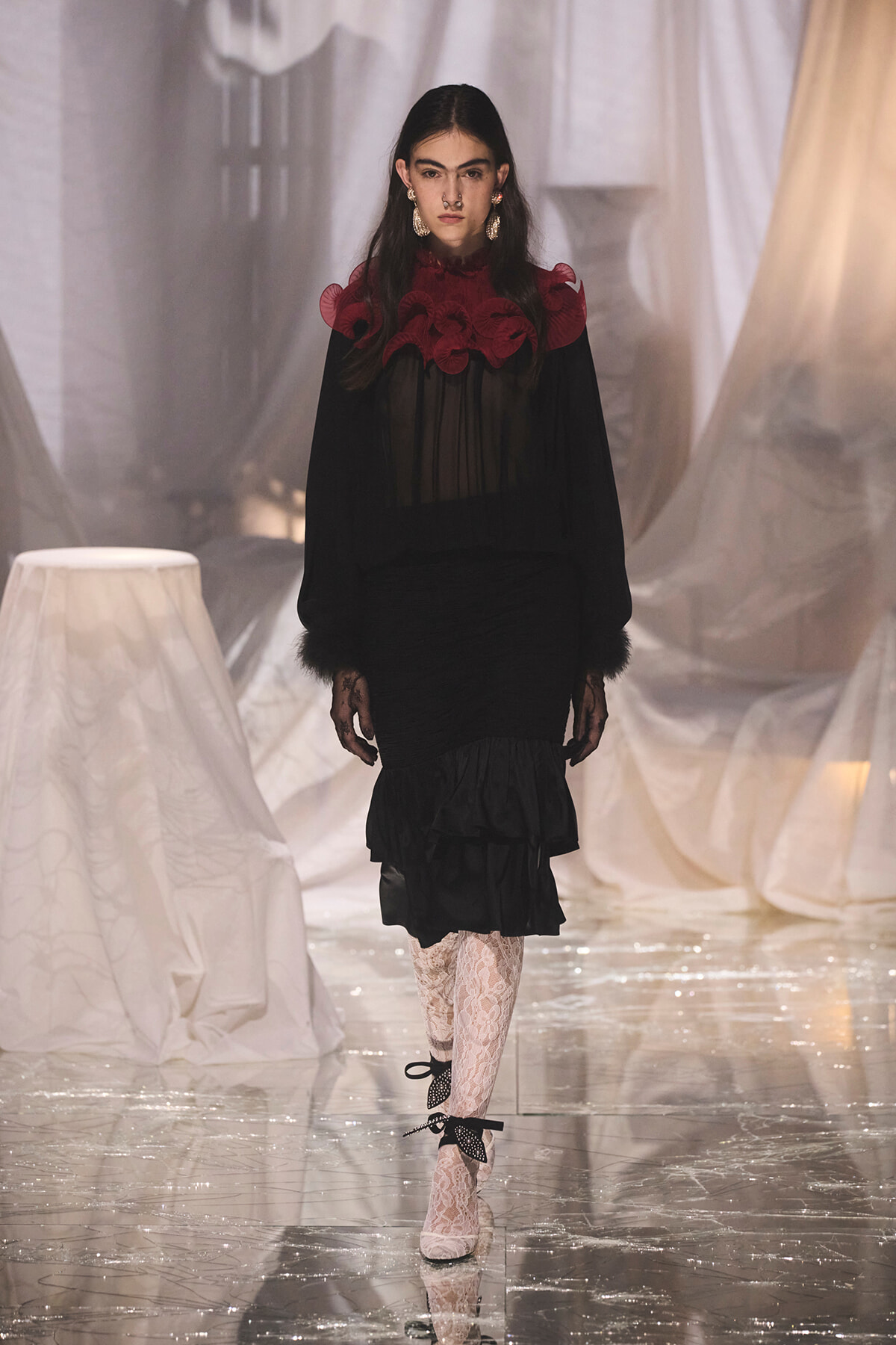 Model on a fashion runway wearing a black sheer blouse with a red ruffle collar, black skirt, lace gloves, and bow-strap heels with lace tights.