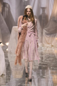 Model walks a fashion runway in a pale pink striped dress with a blush fur stole, lace tights, and a cream knit cap, accessorized with chunky jewelry