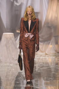 Female model walks the runway in a brown polka-dot suit with black lapels and a pink satin bow at the waist.