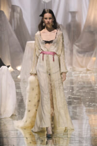 Model on a fashion runway wearing a cream lace gown with a pink belt and a fur stole draped over her arm.