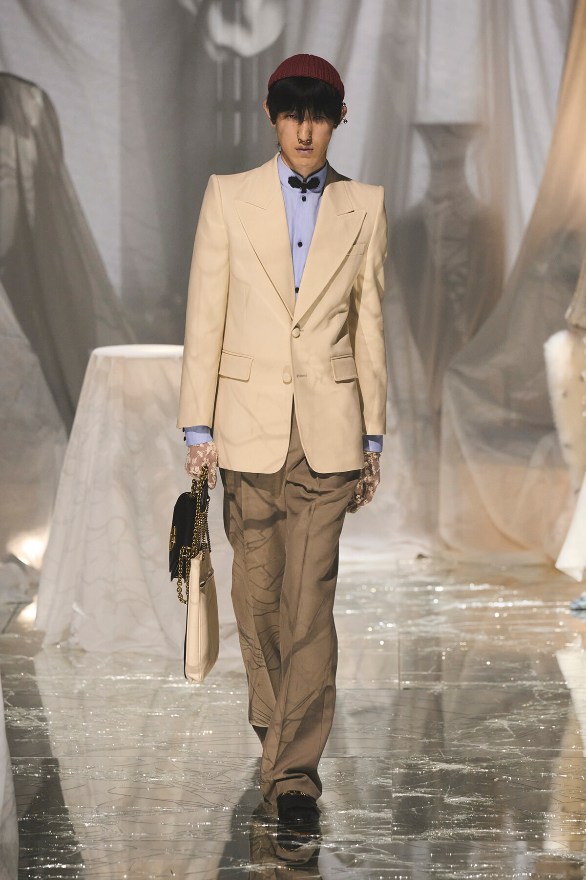 Model walking a runway in a beige suit with a light blue shirt, brown trousers, red beanie, lace gloves, and a beige handbag.