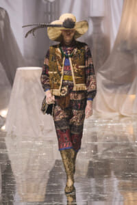 Model on a runway wearing an ornate patterned suit, wide-brim hat with feather, multiple beaded necklaces, gloves, and snakeskin boots.