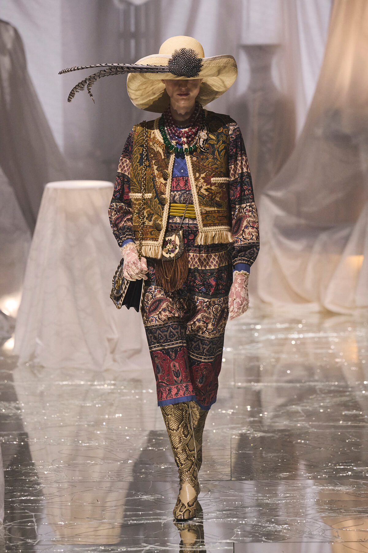 Model on a runway wearing an ornate patterned suit, wide-brim hat with feather, multiple beaded necklaces, gloves, and snakeskin boots.