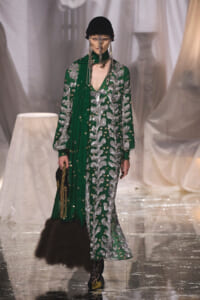 Model on a runway wearing a green gown with silver embroidery, a black knit cap, dangling chains, and a fur-trimmed bag.