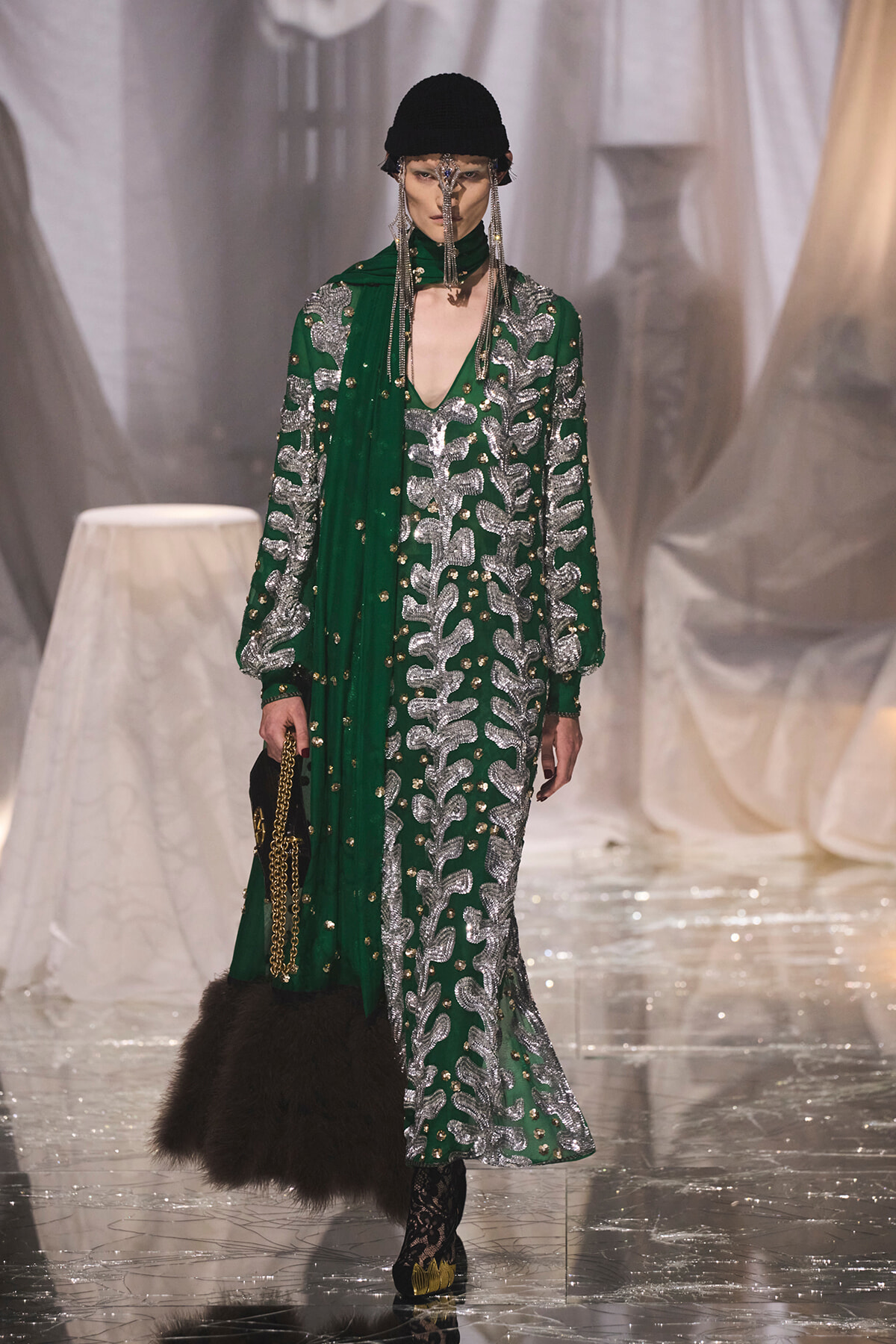 Model on a runway wearing a green gown with silver embroidery, a black knit cap, dangling chains, and a fur-trimmed bag.
