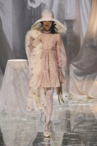 Model walking a fashion runway in a pale pink Victorian-inspired dress with lace trim and sheer sleeves, paired with a wide-brim hat and fur stole.