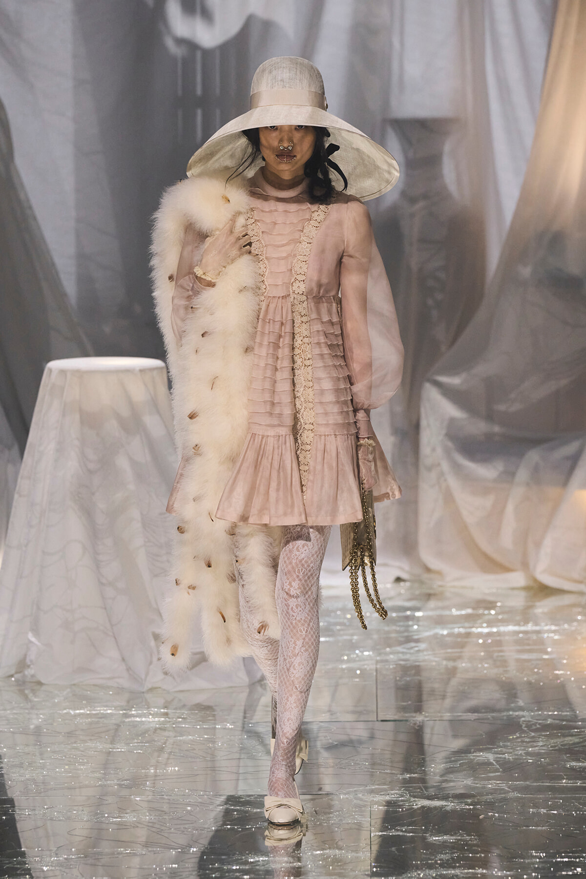 Model walking a fashion runway in a pale pink Victorian-inspired dress with lace trim and sheer sleeves, paired with a wide-brim hat and fur stole.