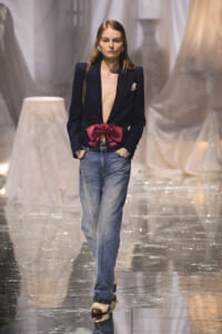 Model on a fashion runway wearing a navy blazer with a deep plunging front and a pink satin bow at the waist, paired with relaxed jeans.
