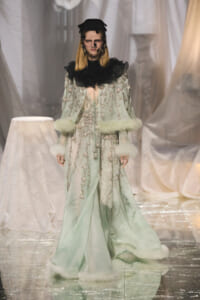 Model walks a runway in a mint-green gown with silver beading, black ruffled collar, and white fur trim at cuffs and hem.