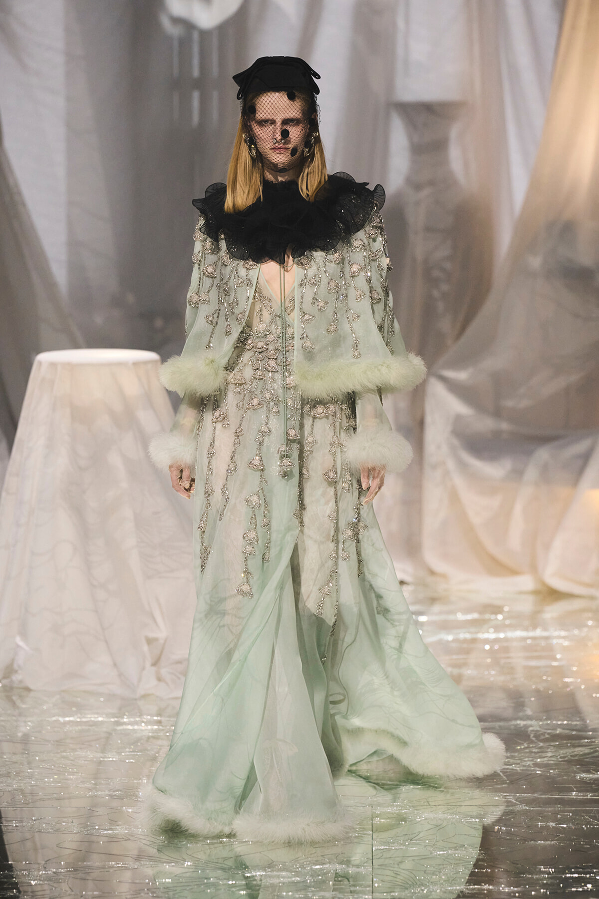 Model walks a runway in a mint-green gown with silver beading, black ruffled collar, and white fur trim at cuffs and hem.