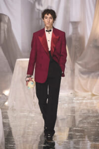 Male model on a runway wearing a burgundy tuxedo jacket, white shirt, black trousers, and a black bow tie, holding a pastel clutch.