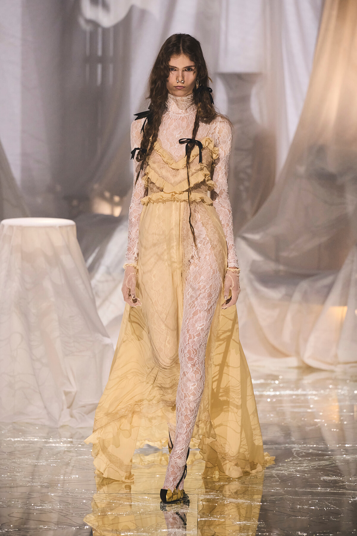 Runway model wearing a pale yellow lace gown with sheer panels, ruffles, and black ribbon accents.