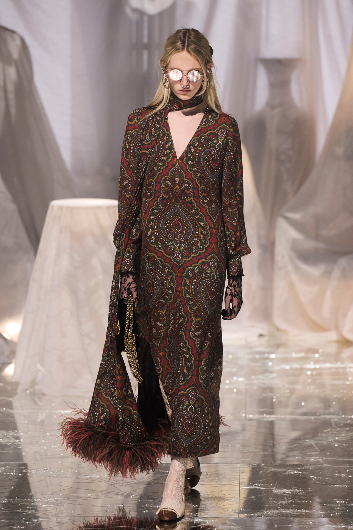 Model walking a fashion runway in a long, burgundy paisley dress with feathered hem and lace gloves, carrying a gold chain handbag.