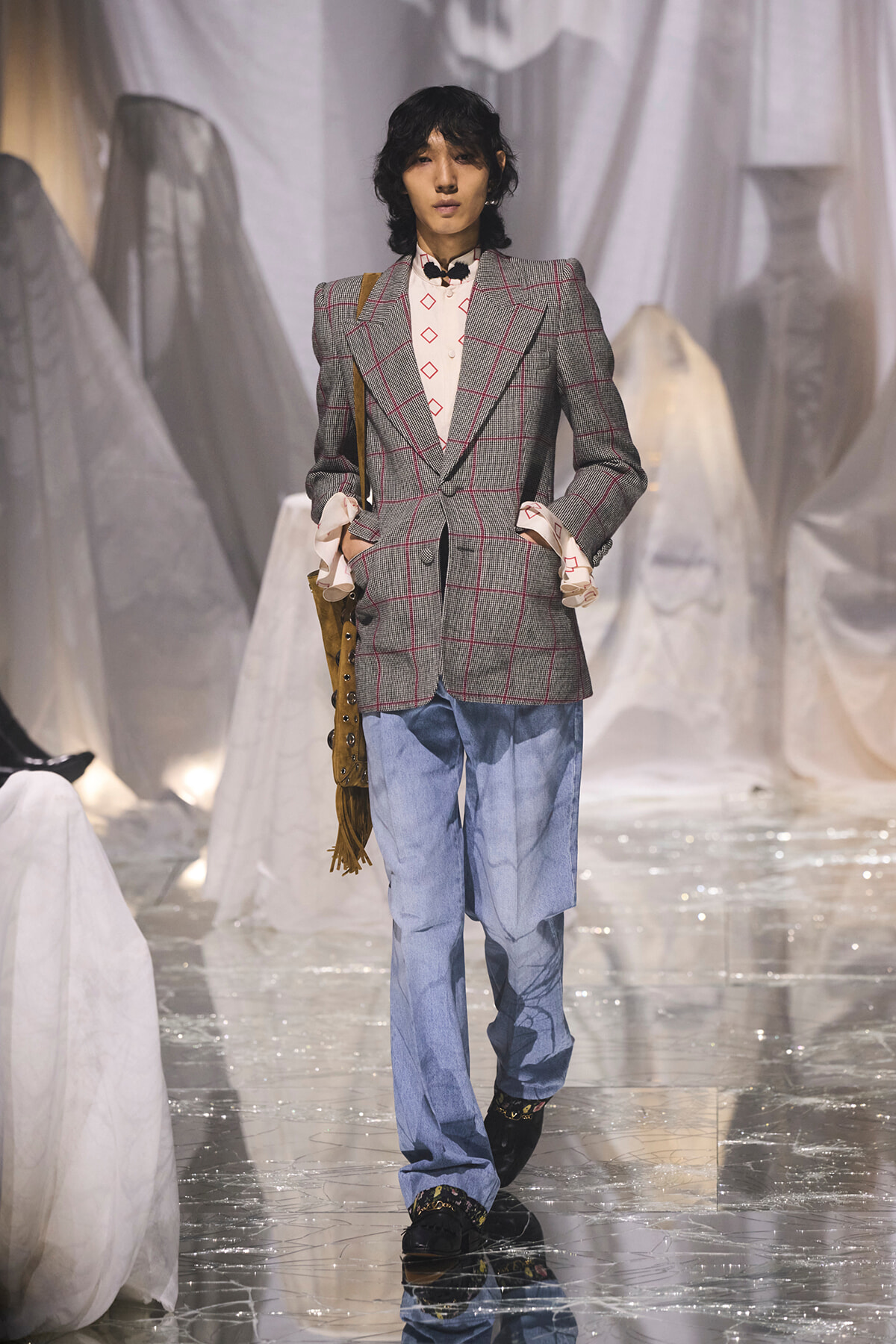 Male model on a fashion runway wearing a grey plaid blazer with red lines, pale pink patterned shirt, loose blue jeans, and a fringed bag over the shoulder.
