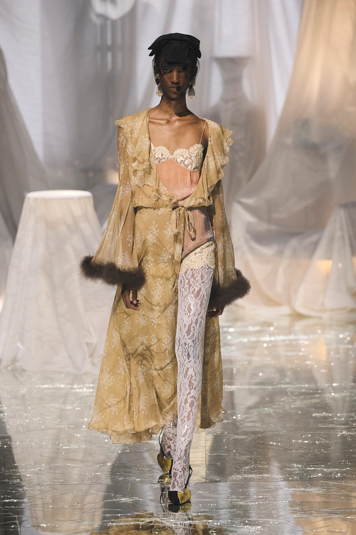 Model walks a fashion runway in a beige floral lace robe over a pink corset, white lace tights, and fur-trimmed cuffs with a black hat and veil.