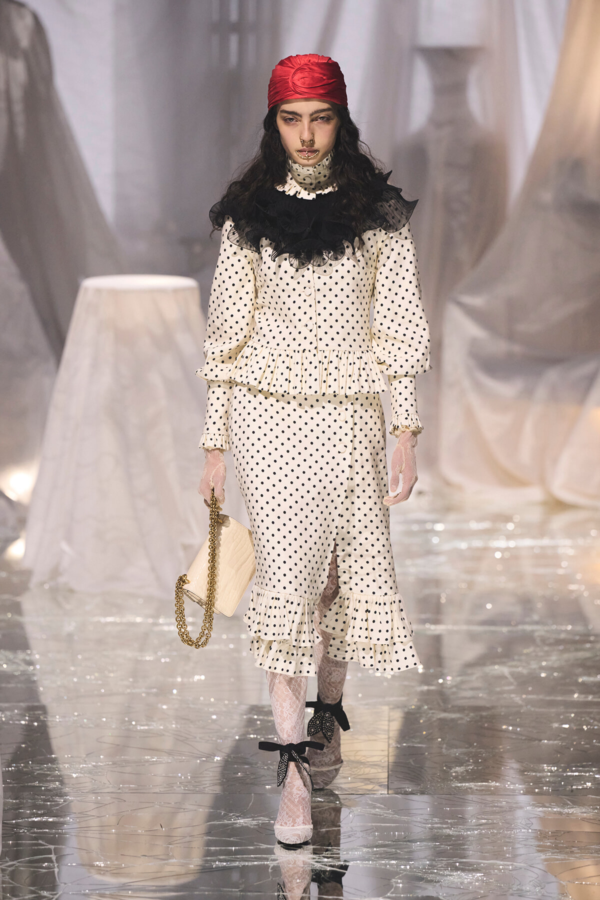 Model walking runway in cream polka-dot dress with ruffled peplum and black lace collar, red headscarf, and lace gloves, carrying a beige clutch with a gold chain template.