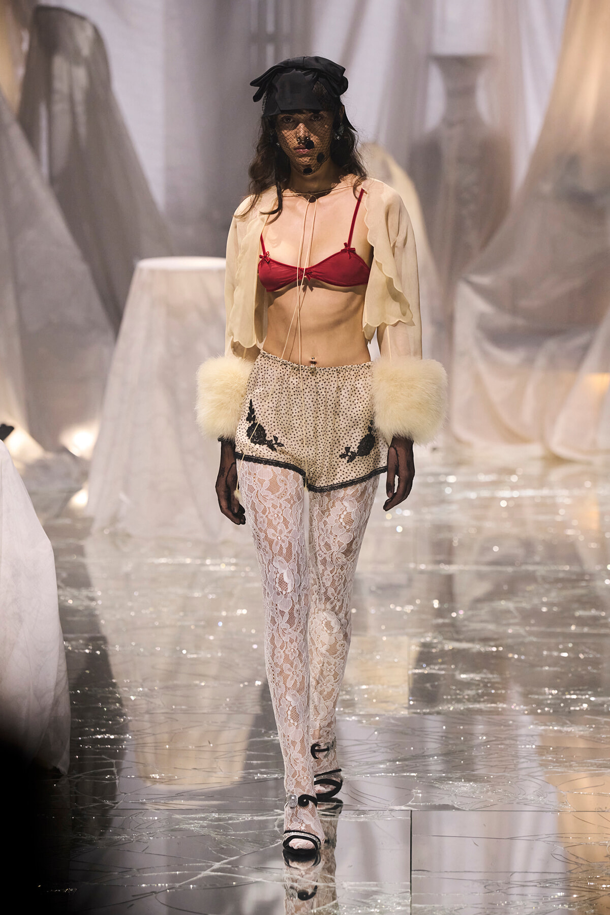 Model on a runway wearing a red bra, sheer beige cape, dotted shorts with black trim, white lace tights, fur cuffs, gloves, and a black veil hat.