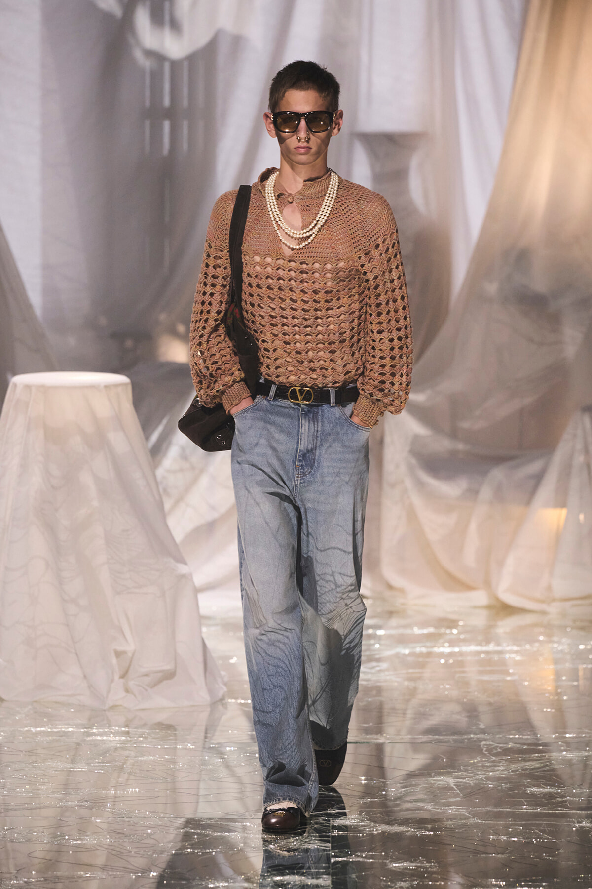 Model on a fashion runway wearing a tan crochet sweater, layered pearl necklaces, light-blue wide-leg jeans, a black belt, sunglasses, and a black shoulder bag.