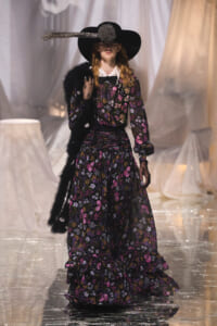 Model on a runway wearing a black floral maxi dress, wide-brim hat with a feather, and black gloves.
