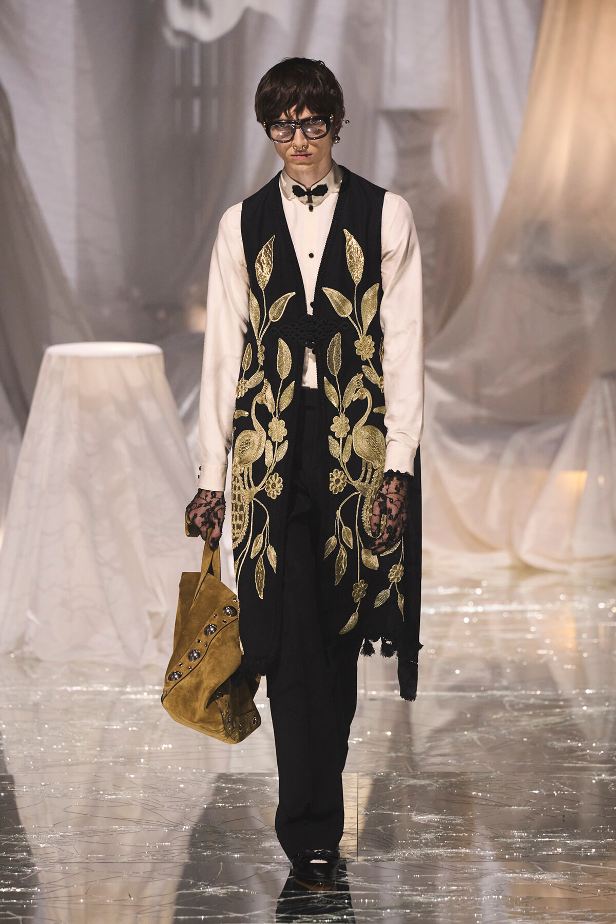 Model on a runway wearing a white shirt and black embroidered vest with gold floral patterns, carrying a tan bag.