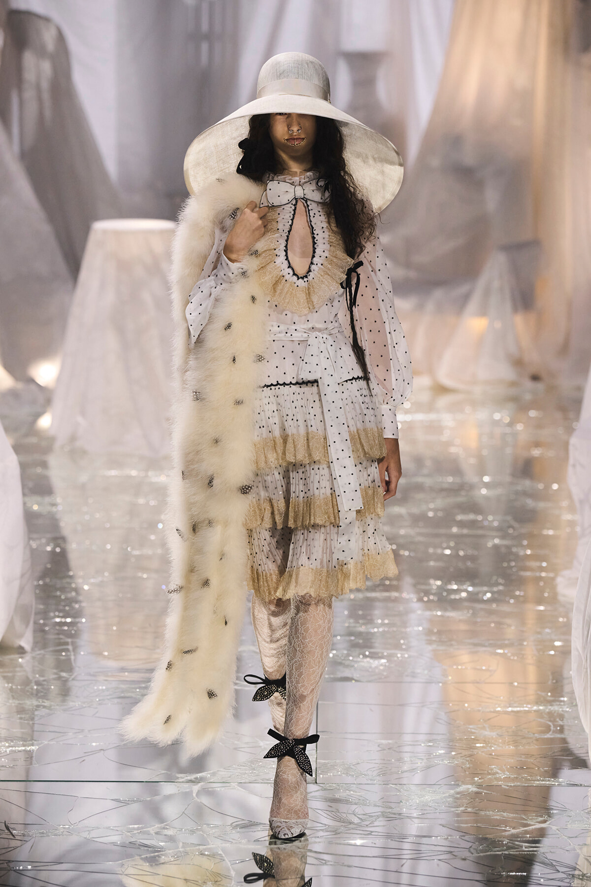 Model in an oversized white hat, polka-dot dress with ruffles, and a fur stole on a glossy runway.