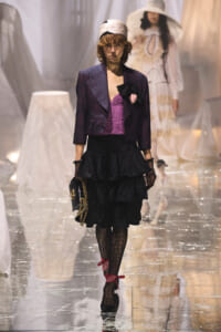 Model on a fashion runway wearing a beige headscarf, navy polka-dot cropped blazer, purple corset, black ruffled skirt, lace tights, and a black handbag with a gold chain.