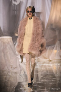 Model walks a glossy runway in a pale beige dress with a oversized pink fur coat, lace tights, round sunglasses, and a large black bow headpiece, carrying a chain clutch.
