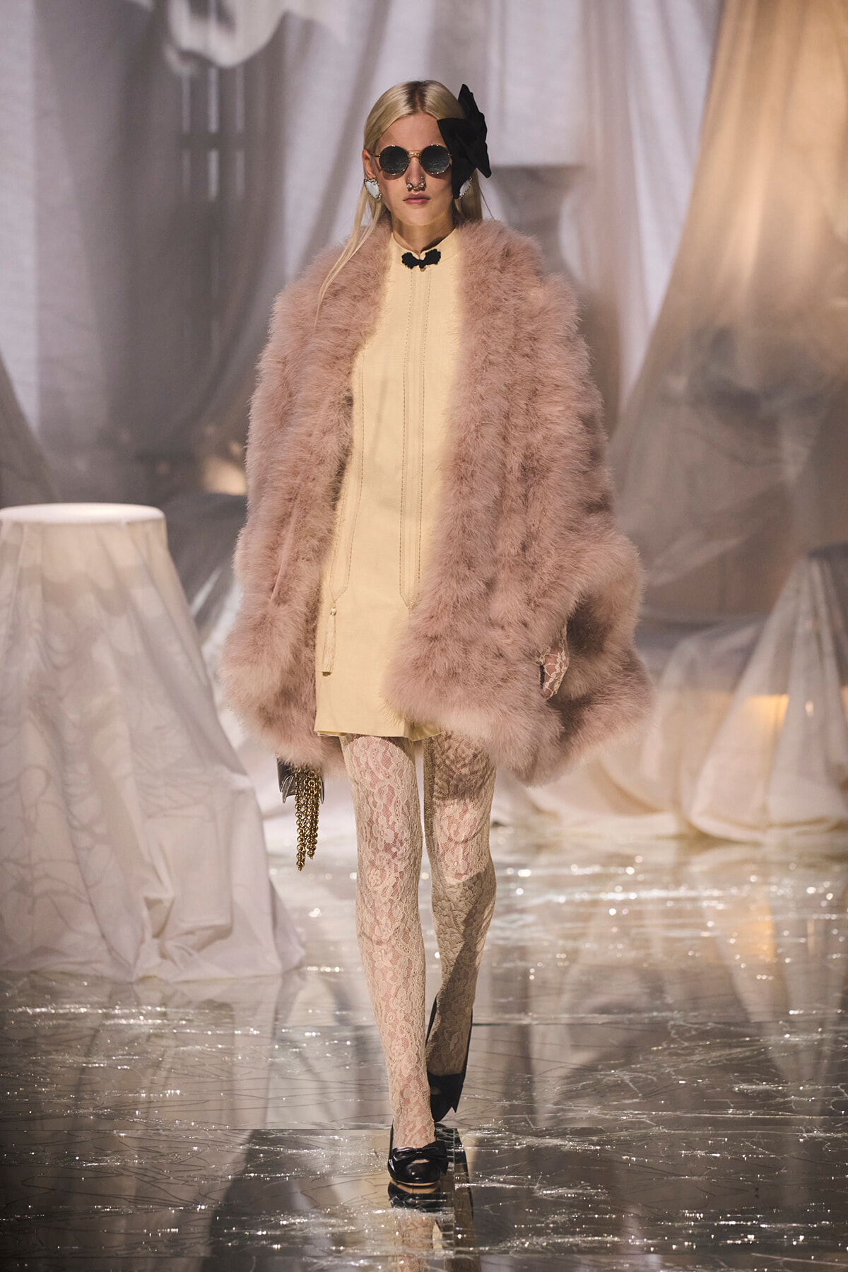 Model walks a glossy runway in a pale beige dress with a oversized pink fur coat, lace tights, round sunglasses, and a large black bow headpiece, carrying a chain clutch.