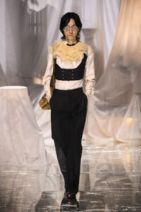 Model on a glossy runway wearing a cream blouse with a beige ruffled collar, a black corset-style vest, and high-waisted black trousers, plus round sunglasses and a brown bag across her body.