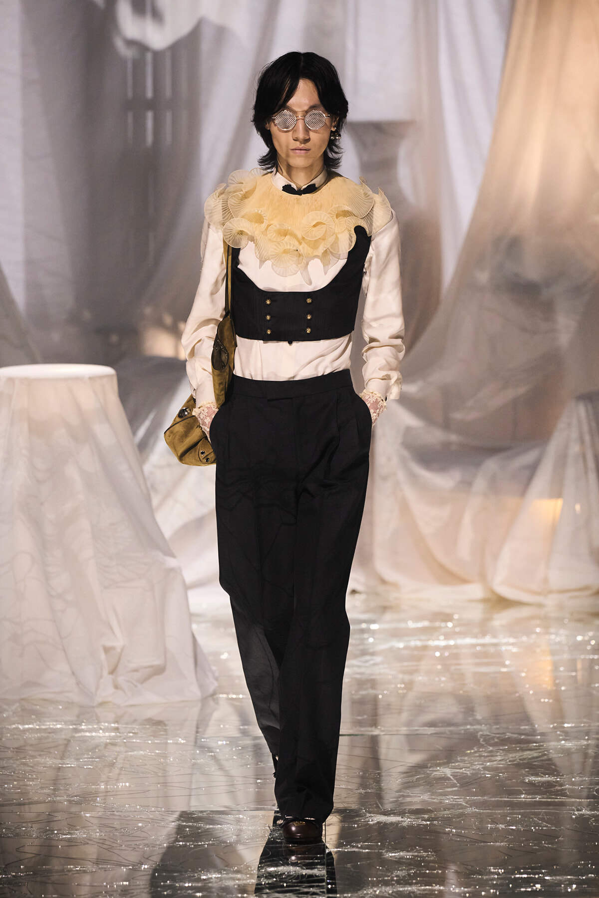Model on a glossy runway wearing a cream blouse with a beige ruffled collar, a black corset-style vest, and high-waisted black trousers, plus round sunglasses and a brown bag across her body.